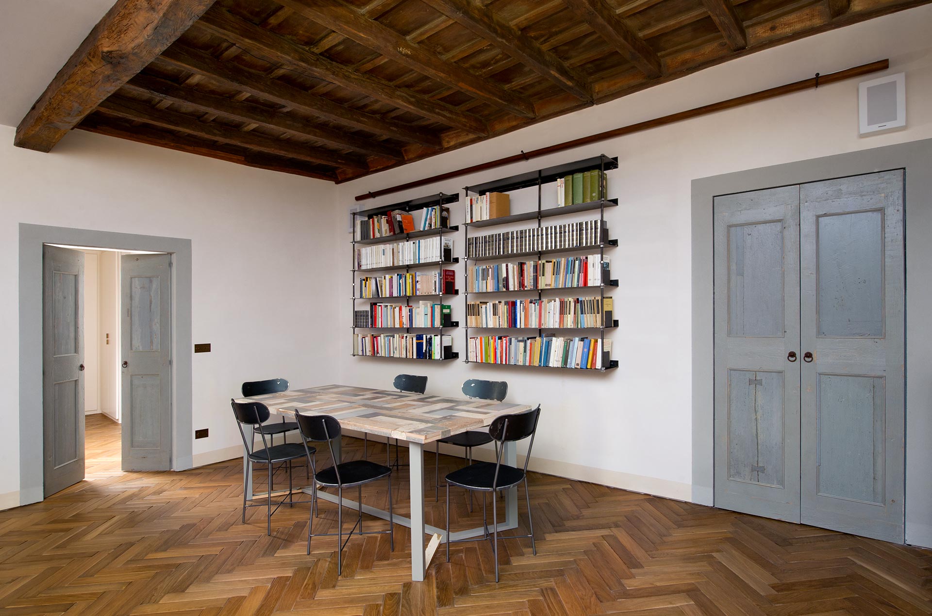 Soggiorno con parquet a spina in legno naturale e grande tavolo in legno colorato nel centro. Ai lati della stanza porte a battenti in legno antico grigio carta da zucchero