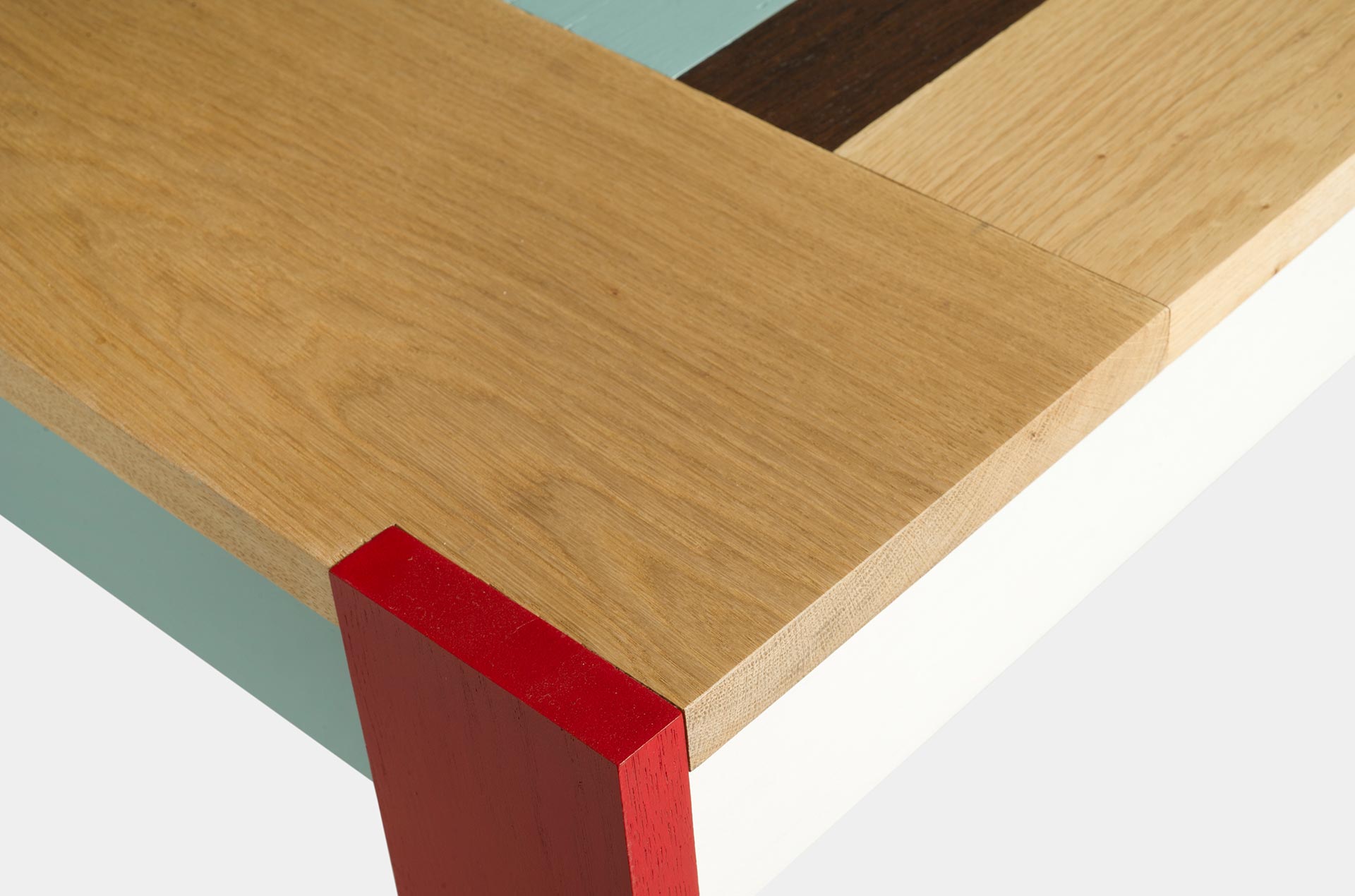 Primo piano di un piano di tavolo artigianale con un mix di legni nuovi e riciclati, colorati e al naturale. Una selle gambe è rossa e si innesta sul piano in rovere