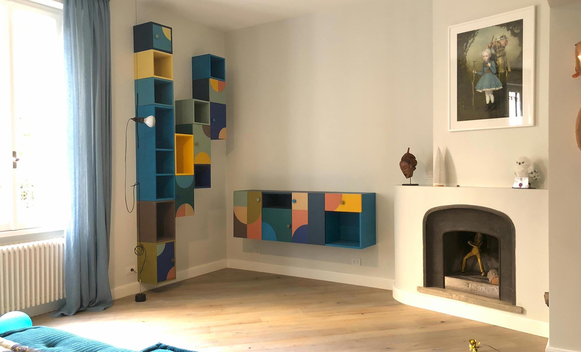 Arredamenti per soggiorno moderno colorati in legno. Il soggiorno presenta un caminetto antico con copertura in marmo bianco e un divano colorato Roche Bobois. Nel fondo parete attrezzata colorata in legno con vani a giorno e vani chiusi da ante.