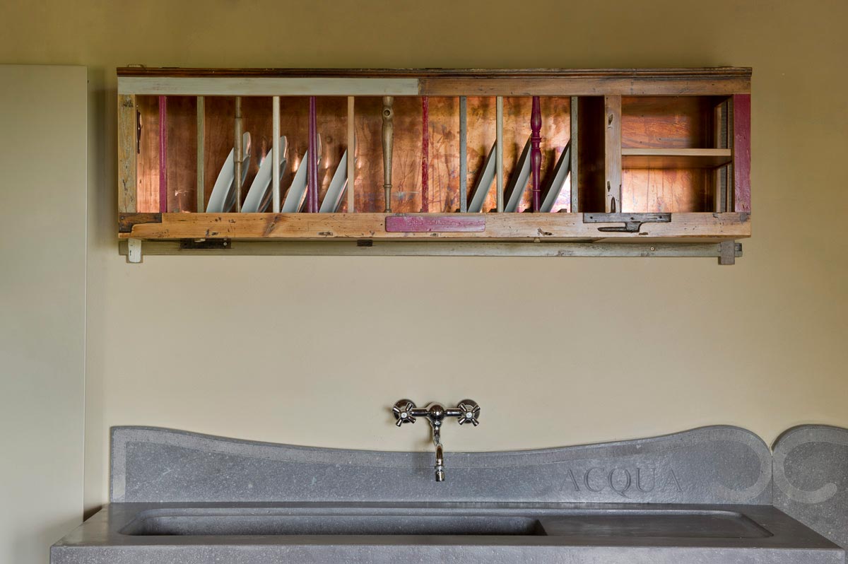 Vista forntale di una piattaia in legno creata a mano con pezzi di legno vintage e vecchie gambe di vecchie sedie in legno colorate. La piattaia è sospesa sopra un antico lavabo in pietra grigio. La schiena della piattaia è in rame nuovo